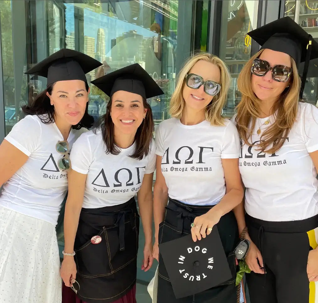 Four women wearing graduation caps and ‘Delta Omega Gamma’ t-shirts; one holds a mortarboard saying ‘IN DOG WE TRUST’, in NYC