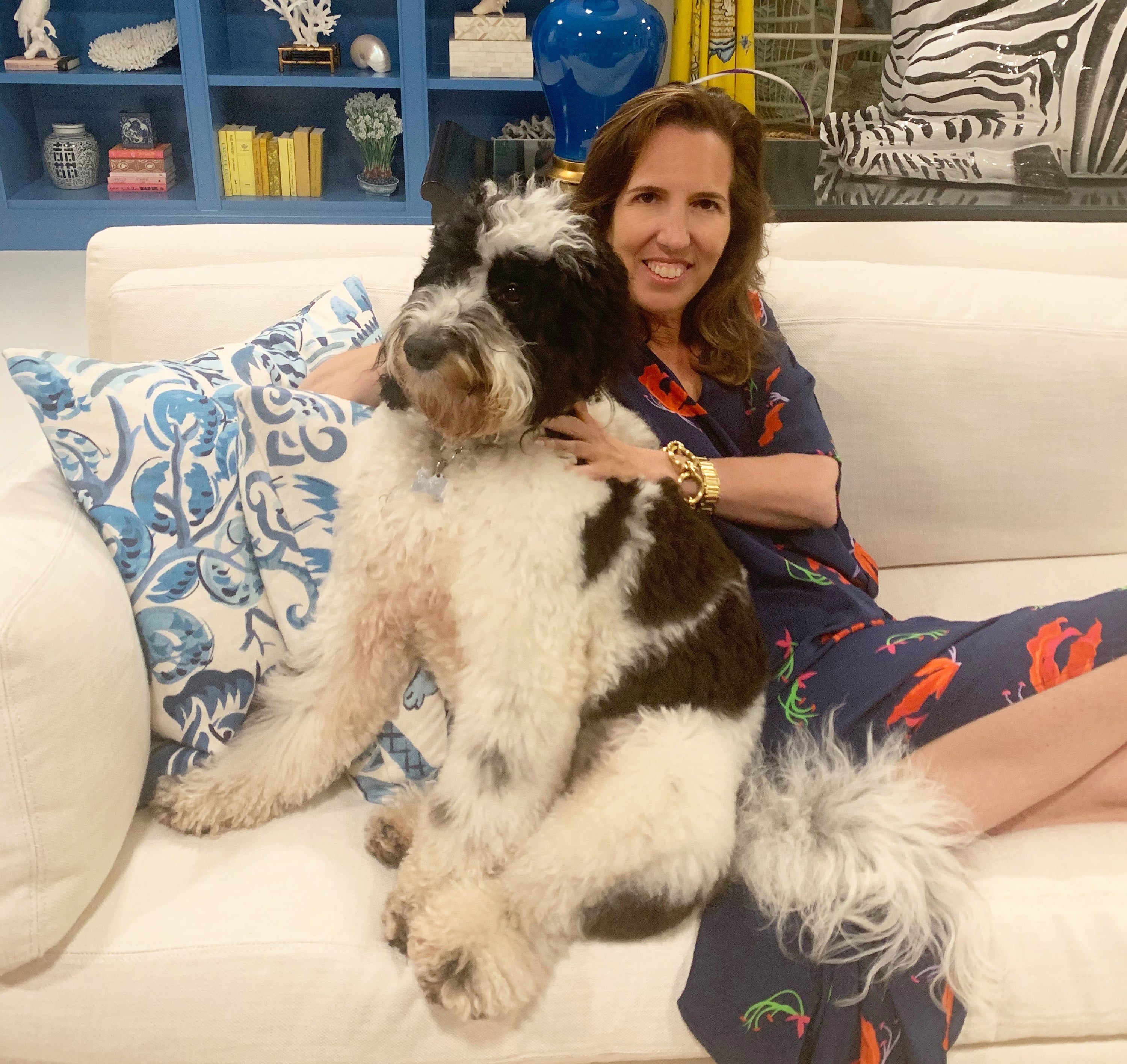 Woman sitting on a white couch hugging a large black-and-white doodle dog in a living room with a blue bookshelf and patterned pillows
