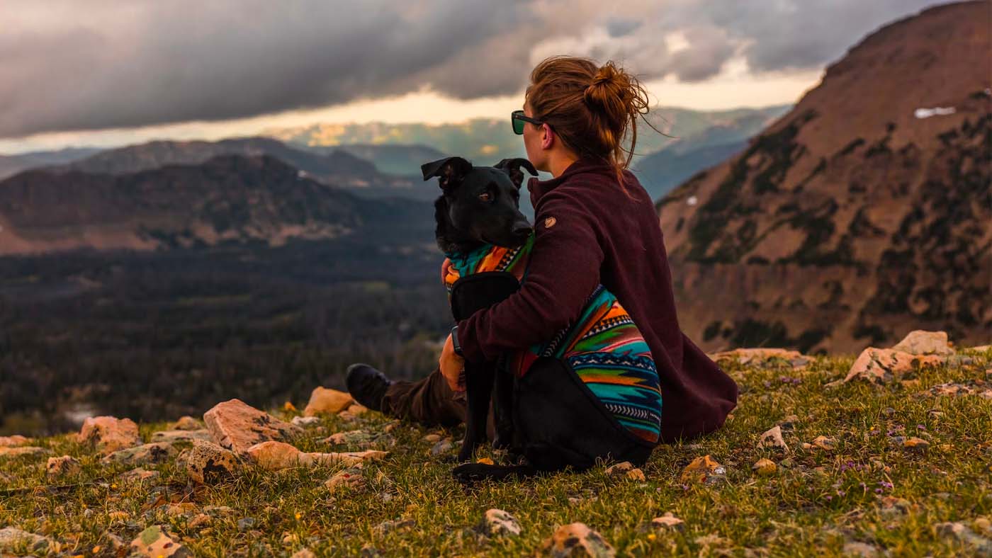 Woman sitting on a mountain ridge holding a black dog in a colorful patterned jacket, overlooking a valley under a cloudy sky