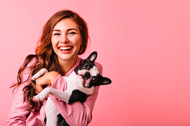 Woman smiling while holding a small black-and-white dog against a pink background