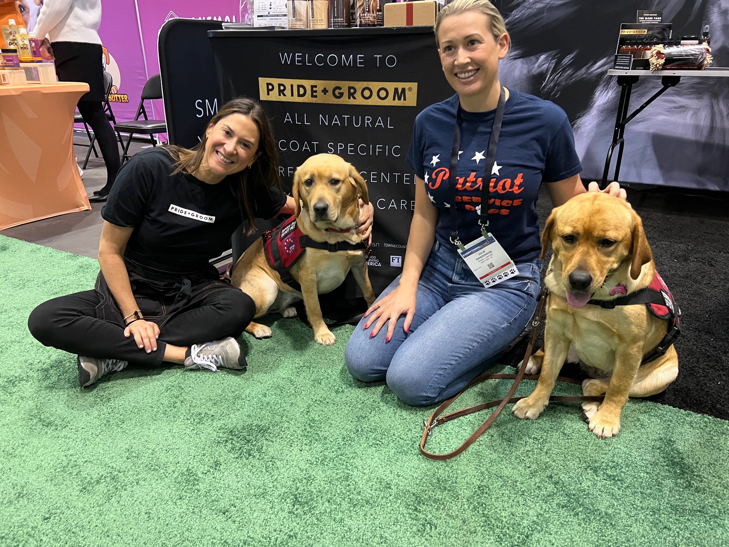 Two women kneeling and smiling while petting two service dogs in red vests in front of a PRIDE+GROOM booth at Global Pet Expo