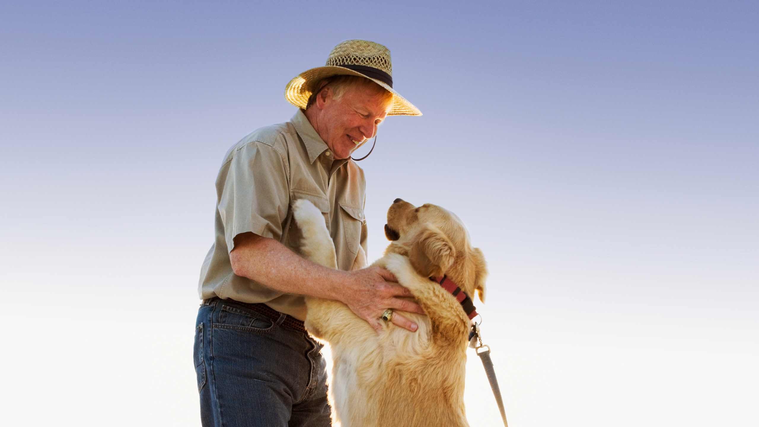 Older man in a straw hat smiling and embracing a golden retriever on a leash against a pale sky