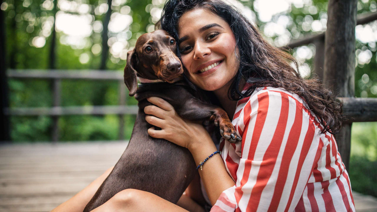 Woman hugging a brown dachshund on a wooden porch with blurred green trees in the background