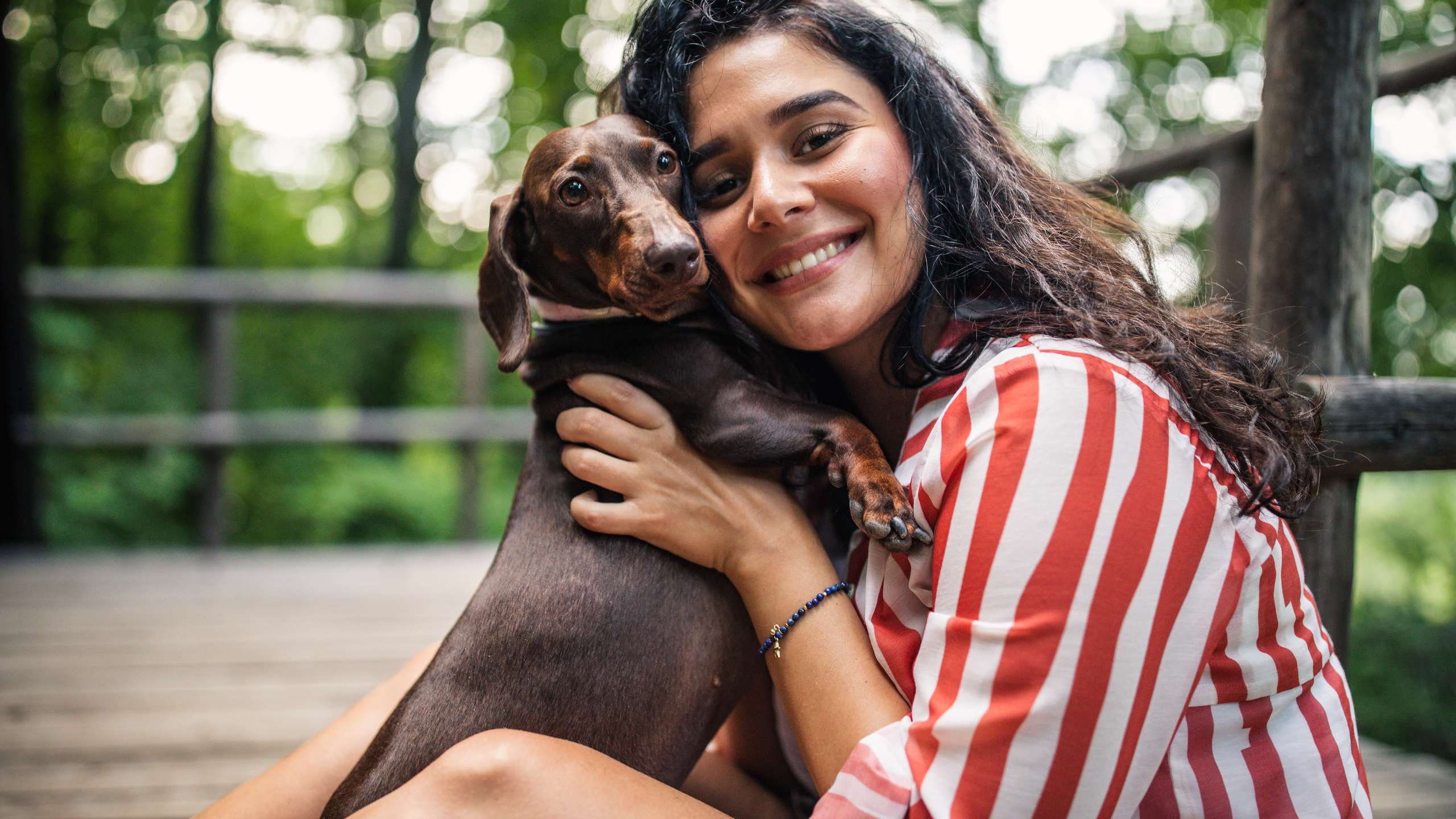 Woman hugging a brown dachshund on a wooden porch with blurred green trees in the background