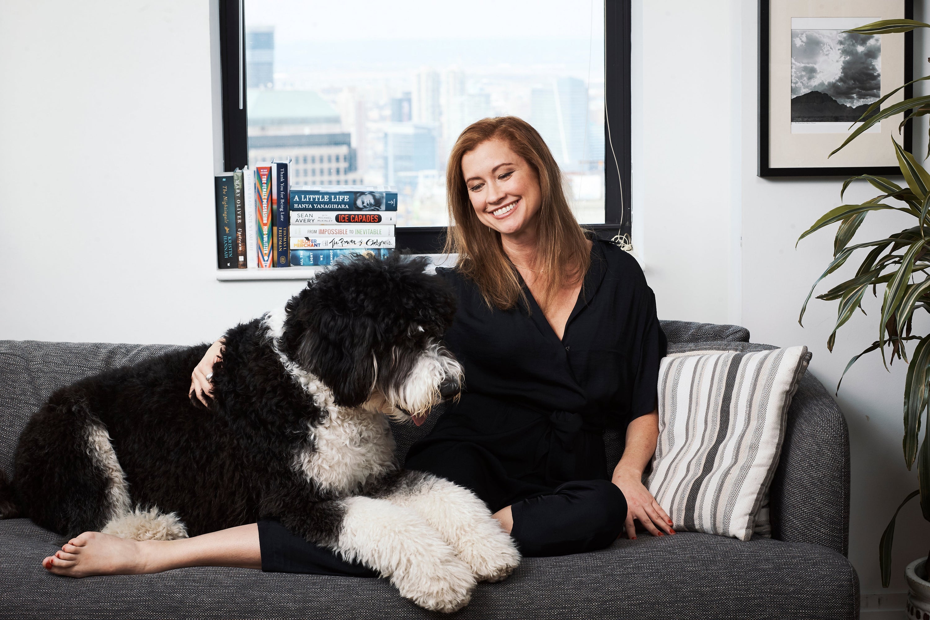 Woman smiling on sofa petting a large black-and-white fluffy dog in an apartment with a city view