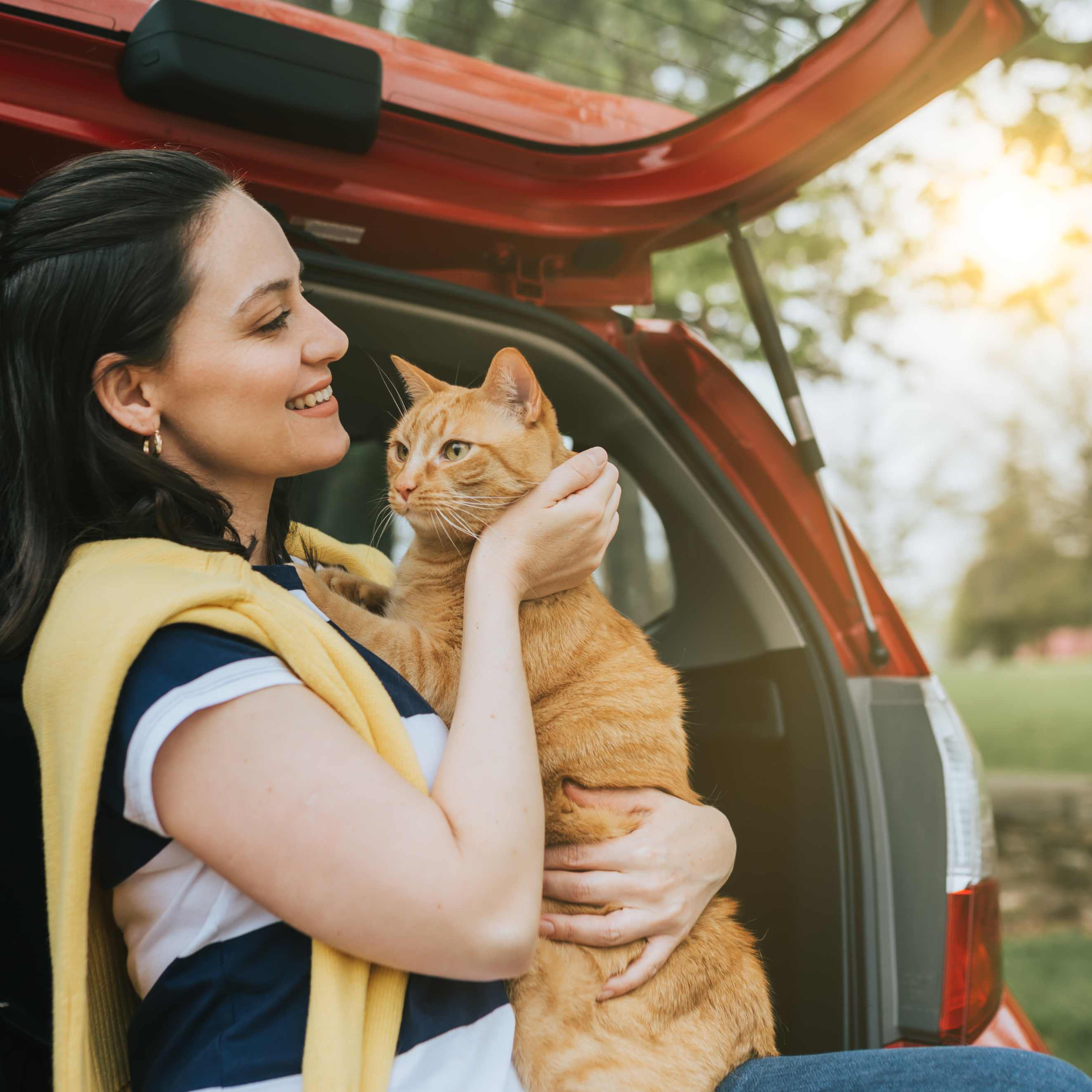 Woman holding an orange tabby cat at the open trunk of a red car, smiling outdoors in soft sunlight