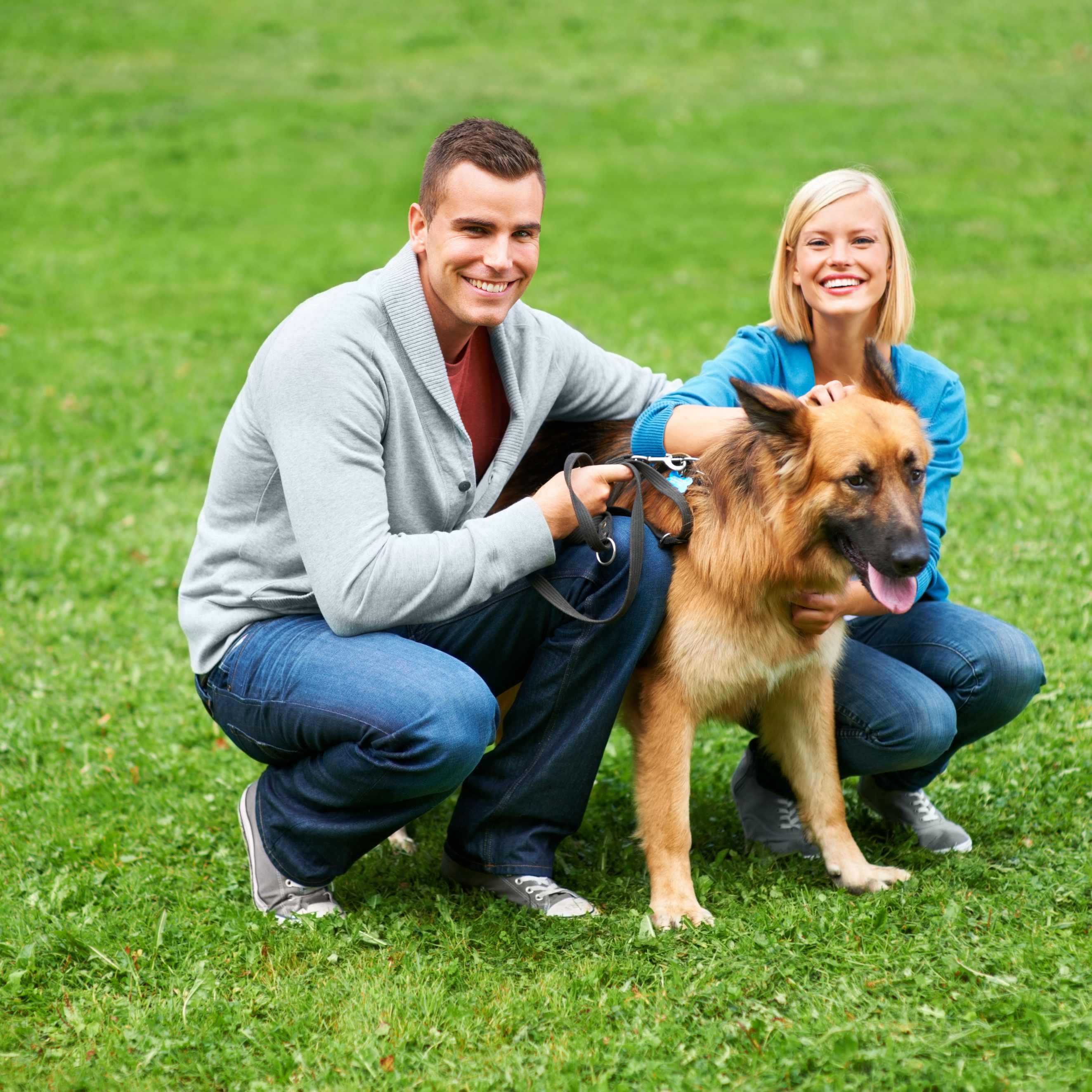 Man and woman crouching and smiling while holding a leashed large brown dog on grass in a park