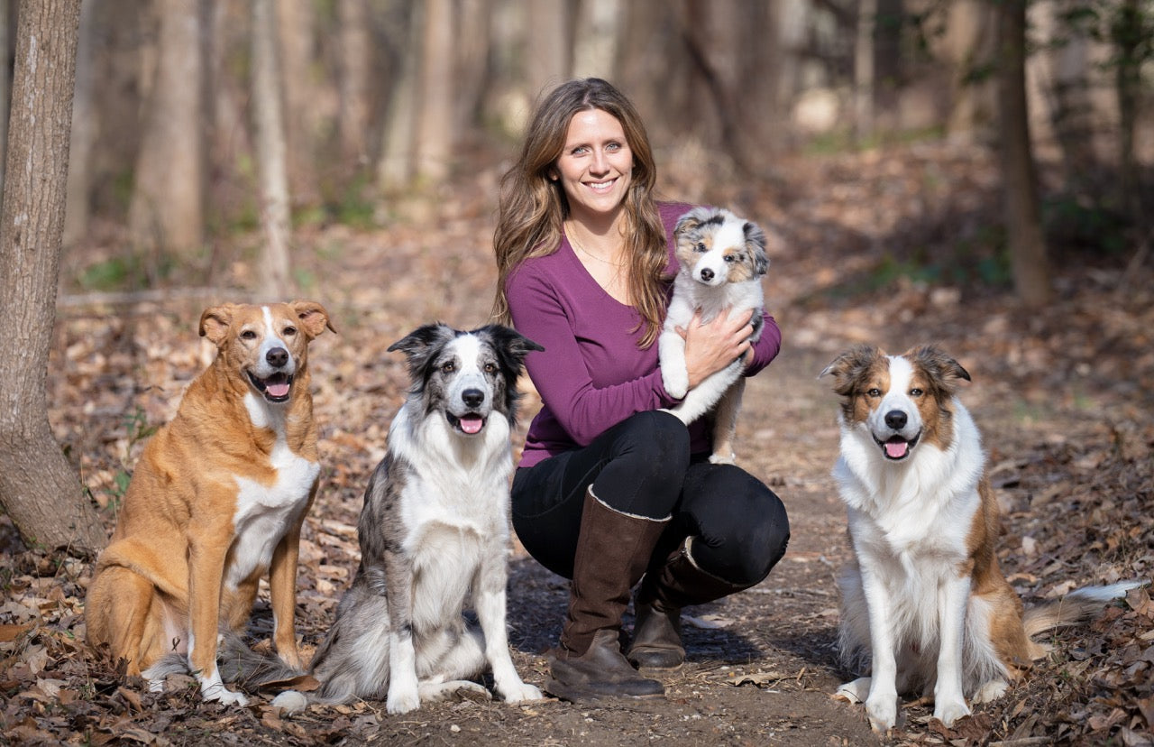 Woman kneeling on a leaf-strewn trail holding a puppy while three dogs sit beside her outdoors