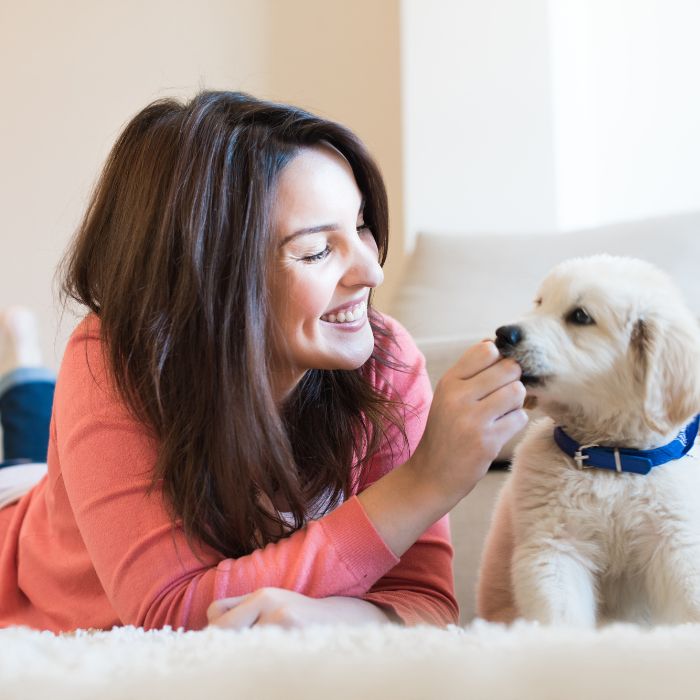 Woman smiling and feeding a light-colored puppy a treat while lying on a white rug in a living room