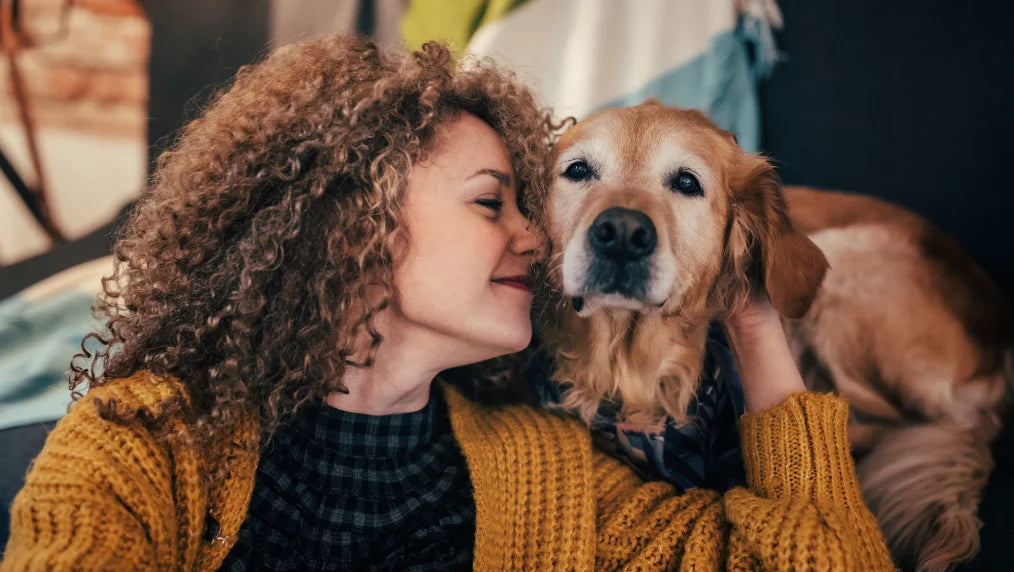 Woman with curly hair cuddling a golden-colored dog on a couch in a warm indoor setting