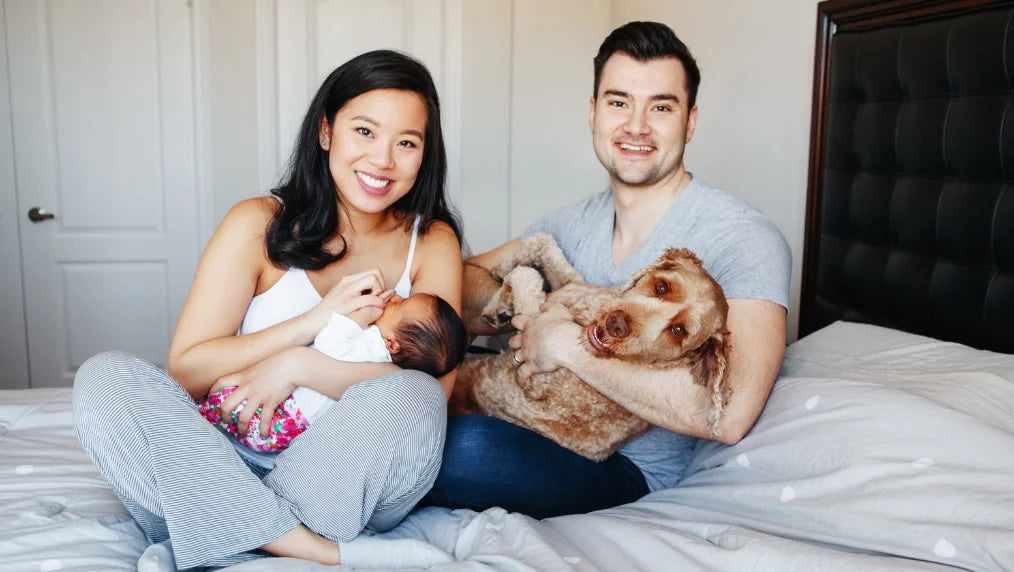 Couple sitting on a bed, mother cradling a newborn while father holds a brown doodle dog in a bright bedroom
