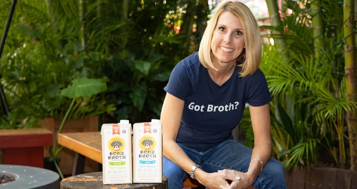 Brutus Broth co-founder seated outdoors smiling beside two bone broth cartons, wearing a navy t-shirt reading Got Broth?