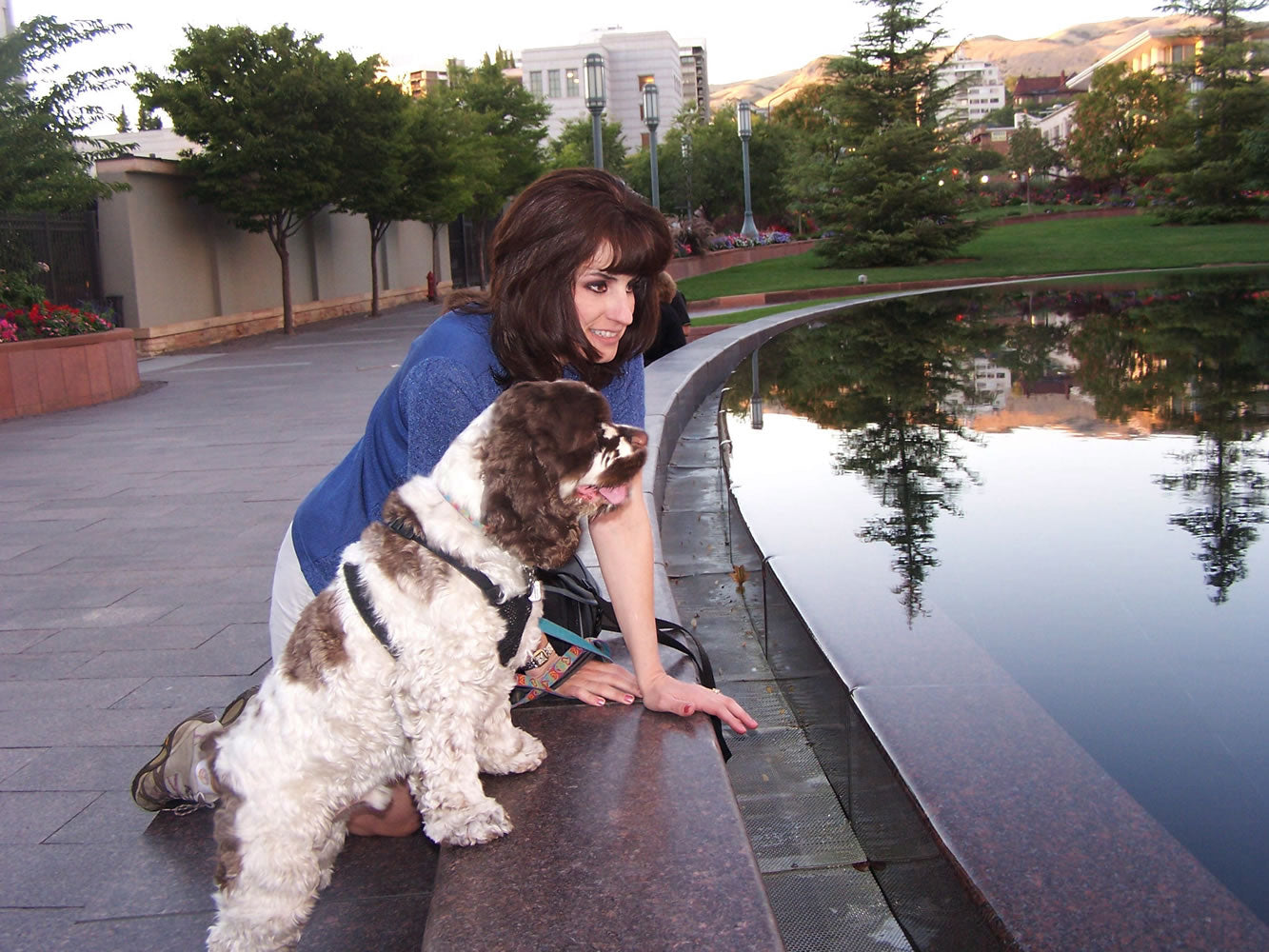 Woman kneeling with a brown-and-white spaniel at the edge of a reflective park fountain