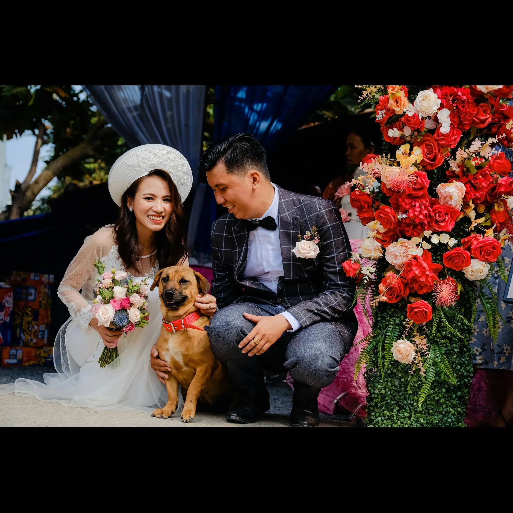 Bride and groom crouch with their dog at an outdoor wedding, bride holding a bouquet beside a tall red and pink floral arrangement