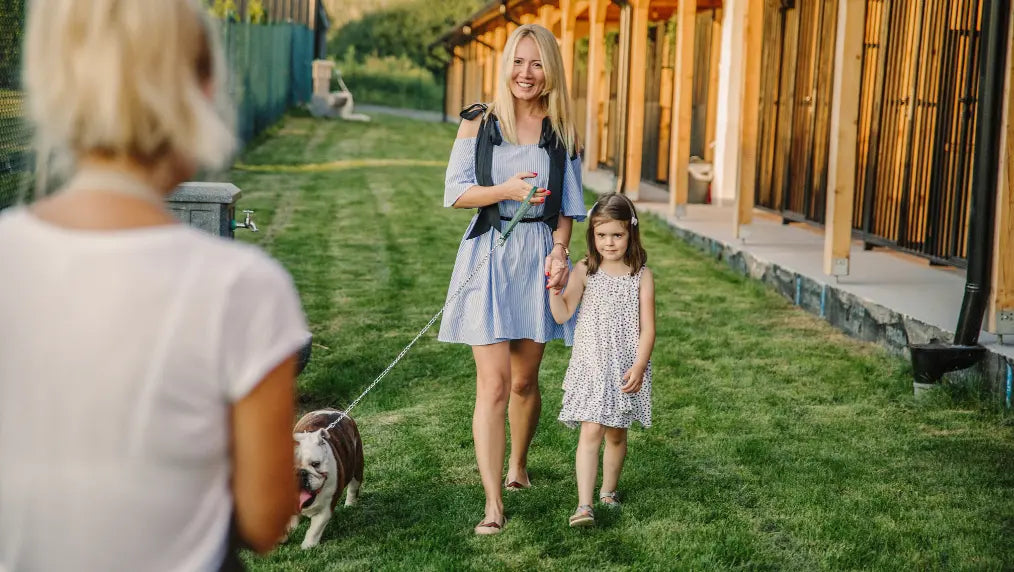 Woman and young girl walking a dog along grass beside kennel buildings while a person watches from the foreground