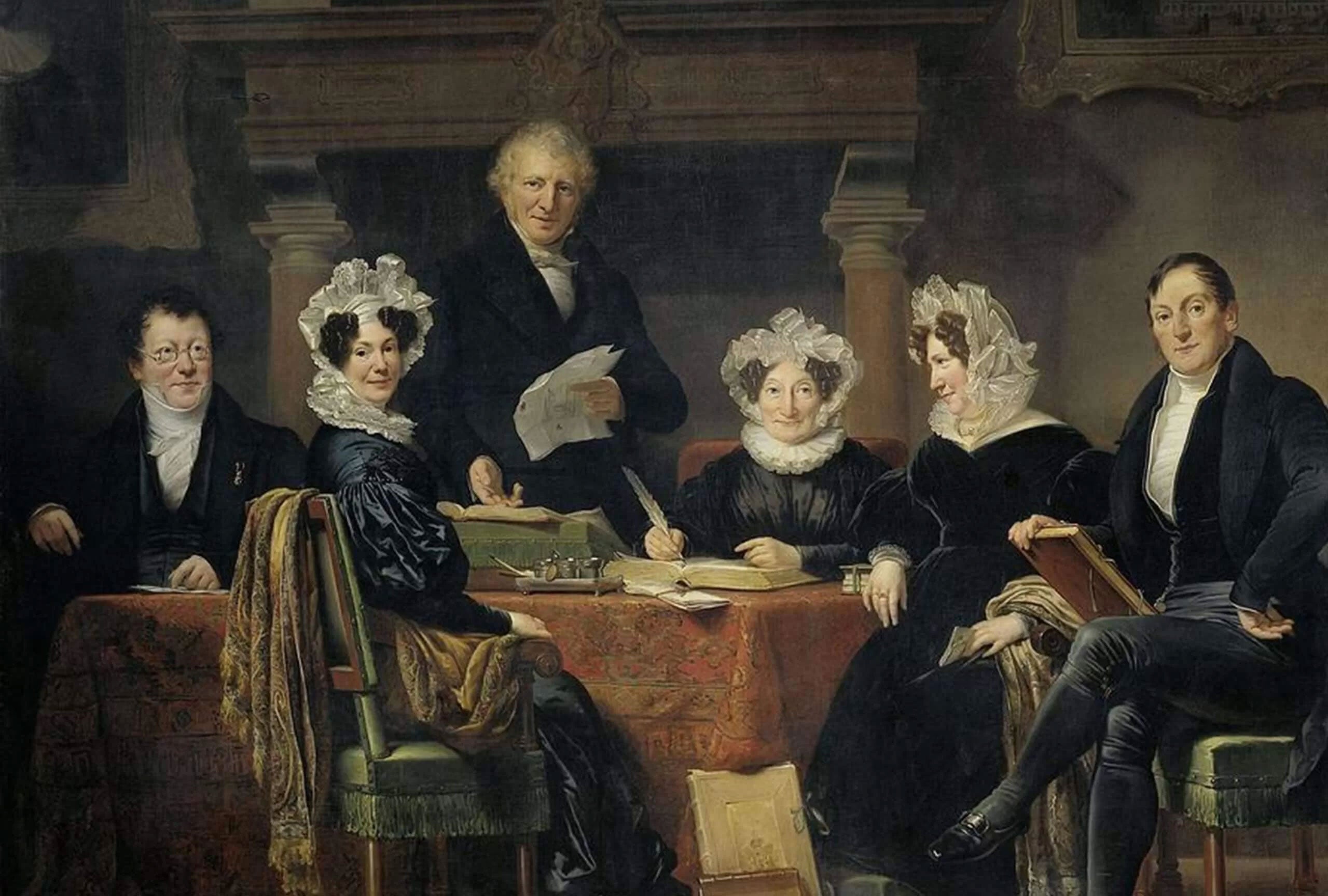Group of six people in early 19th-century dress around a table, one man standing reading papers while others write and listen